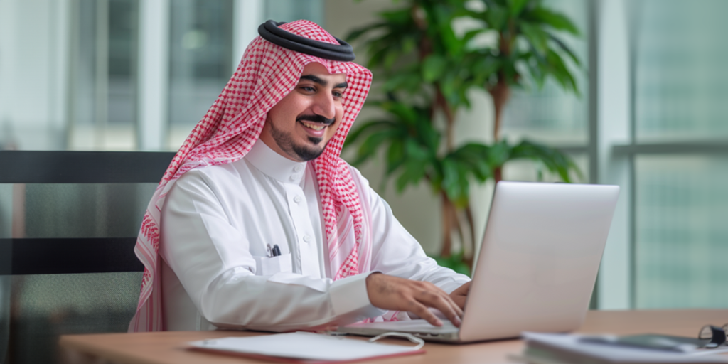 A man wearing a white thobe and red-and-white checkered keffiyeh is sitting at a desk, smiling at his open laptop. He has a pen in his shirt pocket, and a notebook and pen are on the desk. Behind him, a large potted plant and windows with a city view reflect Saudi growth momentum in e-commerce.